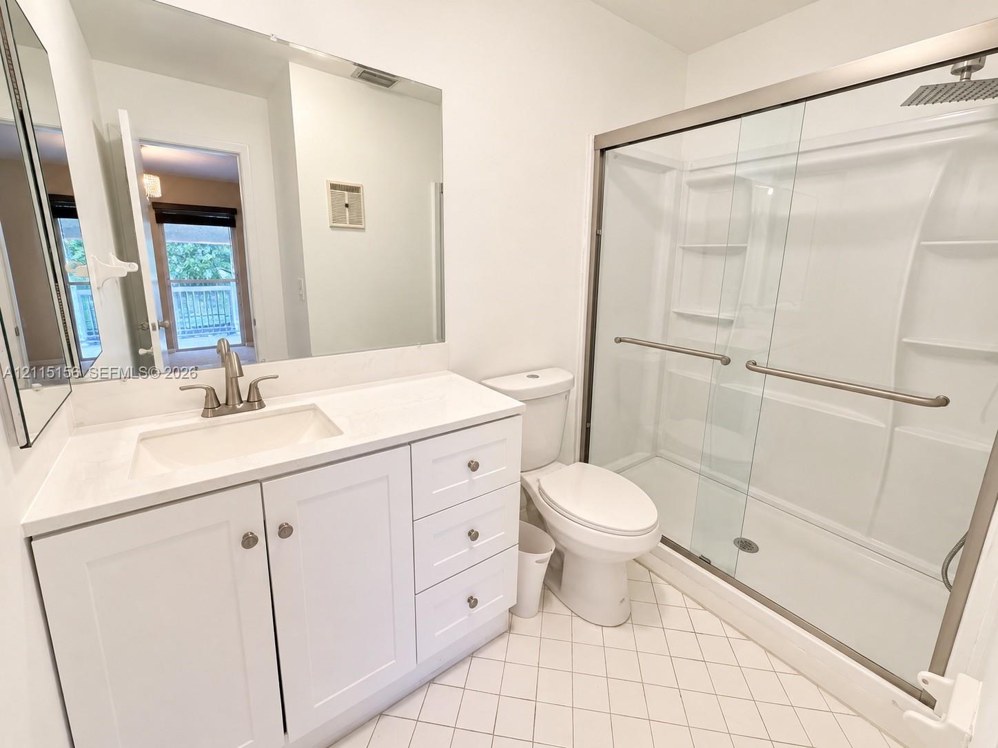 Bathroom featuring white cabinetry, solid-surface countertop, full-width wall mirror, shower with glass sliding doors and integrated shelving, and tiled flooring Bathroom featuring white cabinetry, solid-surface countertop, full-width wall mirror, shower with glass sliding doors and integrated shelving, and tiled flooring