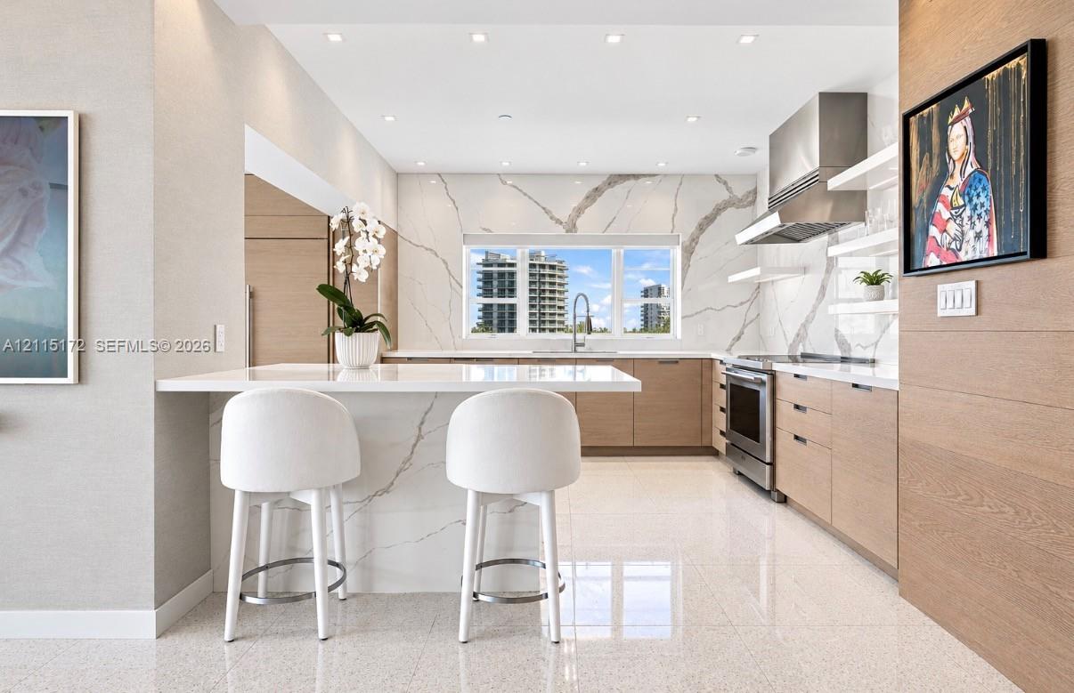 Kitchen featuring a waterfall-edge stone island, veined stone backsplash, stainless steel range hood, flat-panel wood-finish cabinetry, and recessed ceiling lighting