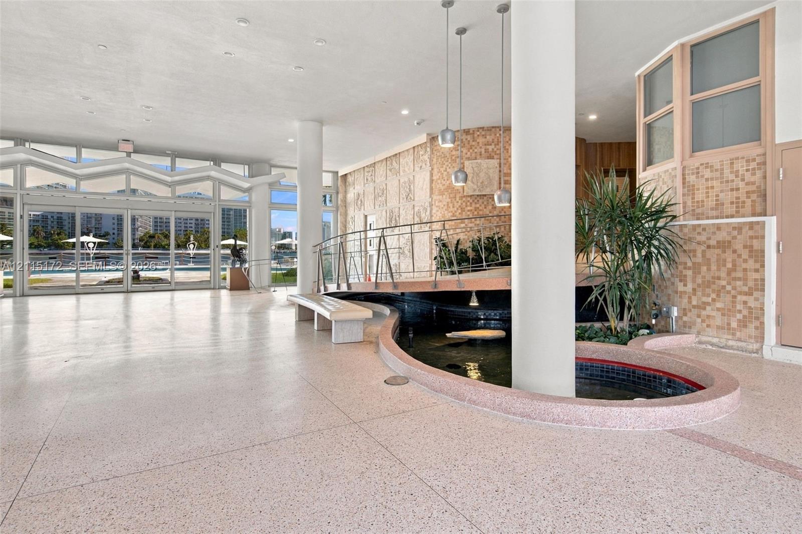Spacious interior featuring terrazzo flooring, a central decorative pond, full-height glass folding doors, structural cylindrical columns, and pendant lighting