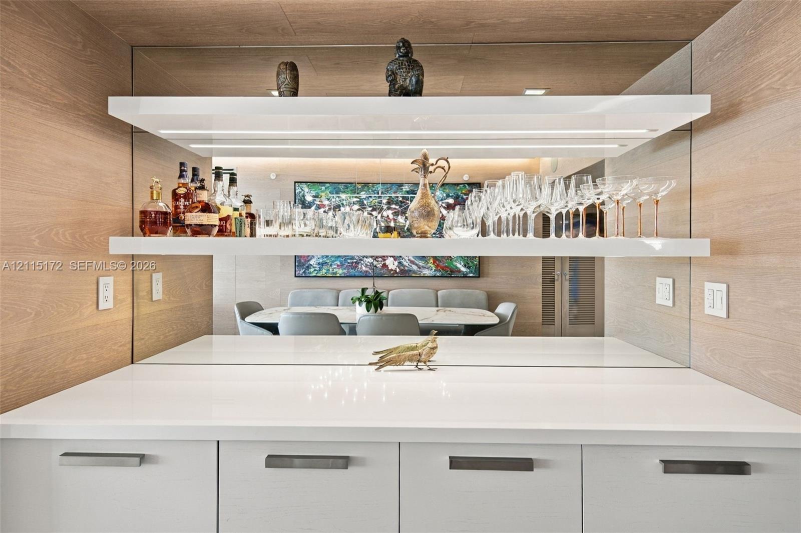 Custom built-in bar featuring white cabinetry, polished solid-surface countertop, mirrored backsplash, and tiered floating shelving with integrated LED lighting, framed by wood-finish wall paneling