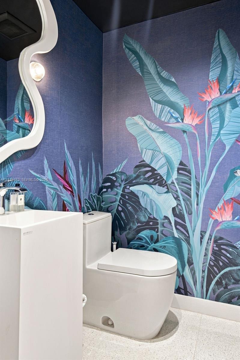 Powder room featuring botanical print wall covering, sculptural white pedestal sink with chrome faucet, high-profile dual-flush toilet, and wall-mounted irregular-shaped mirror