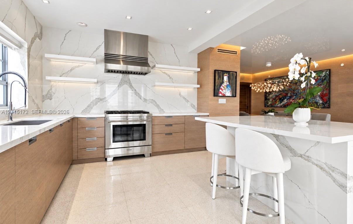 Kitchen featuring flat-panel wood cabinetry, a waterfall-edge island, a floor-to-ceiling stone-slab backsplash, integrated stainless steel range, and floating display shelving