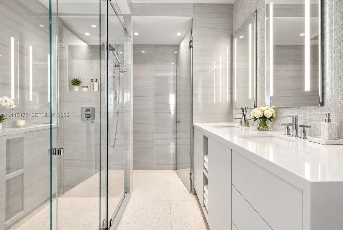 Walk-in glass-enclosed shower featuring large-format tile wall cladding, integrated wall niche, dual-sink vanity with white stone countertop, and vertical LED-lit vanity mirrors