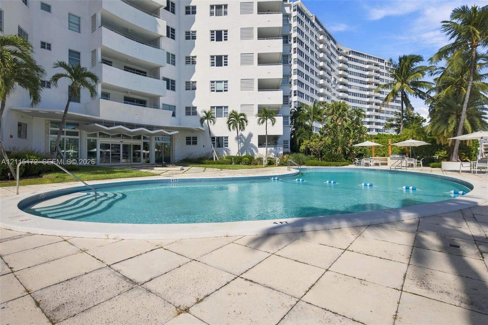 Curved outdoor swimming pool with stone-tile decking, mature palm trees, and ground-level balcony access