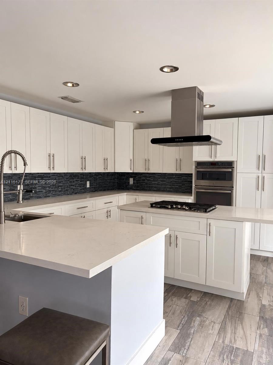 Kitchen featuring white shaker-style cabinetry, quartz countertops, a dark glass tile backsplash, stainless steel built-in double wall ovens with a gas cooktop island, and wood-finish tile flooring Kitchen featuring white shaker-style cabinetry, quartz countertops, a dark glass tile backsplash, stainless steel built-in double wall ovens with a gas cooktop island, and wood-finish tile flooring