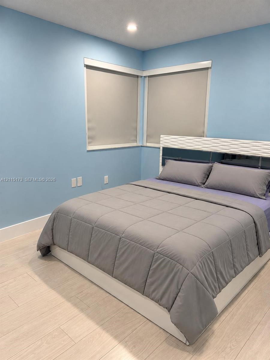 Corner bedroom featuring light-toned wood-finish flooring, recessed ceiling lighting, and dual corner windows with neutral-toned roller shades Corner bedroom featuring light-toned wood-finish flooring, recessed ceiling lighting, and dual corner windows with neutral-toned roller shades