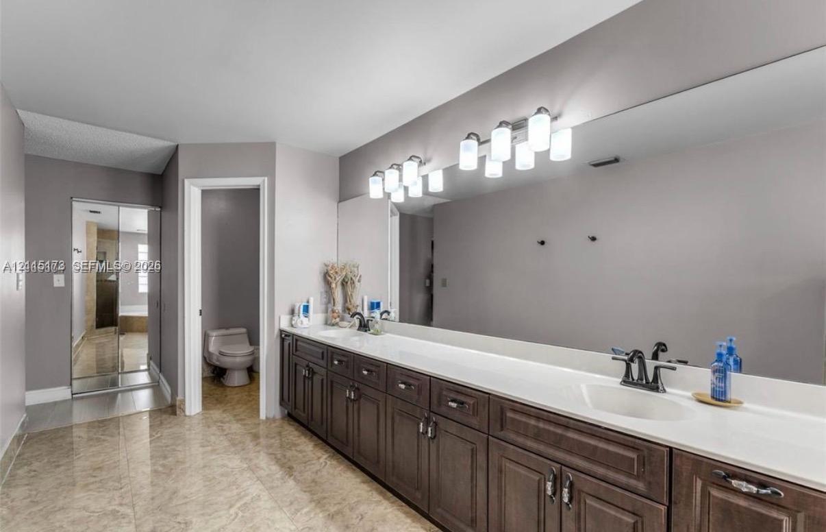 Dual-sink vanity with dark wood cabinetry, integrated white countertops, wide-format wall mirror, multi-bulb overhead lighting, and stone-finish flooring