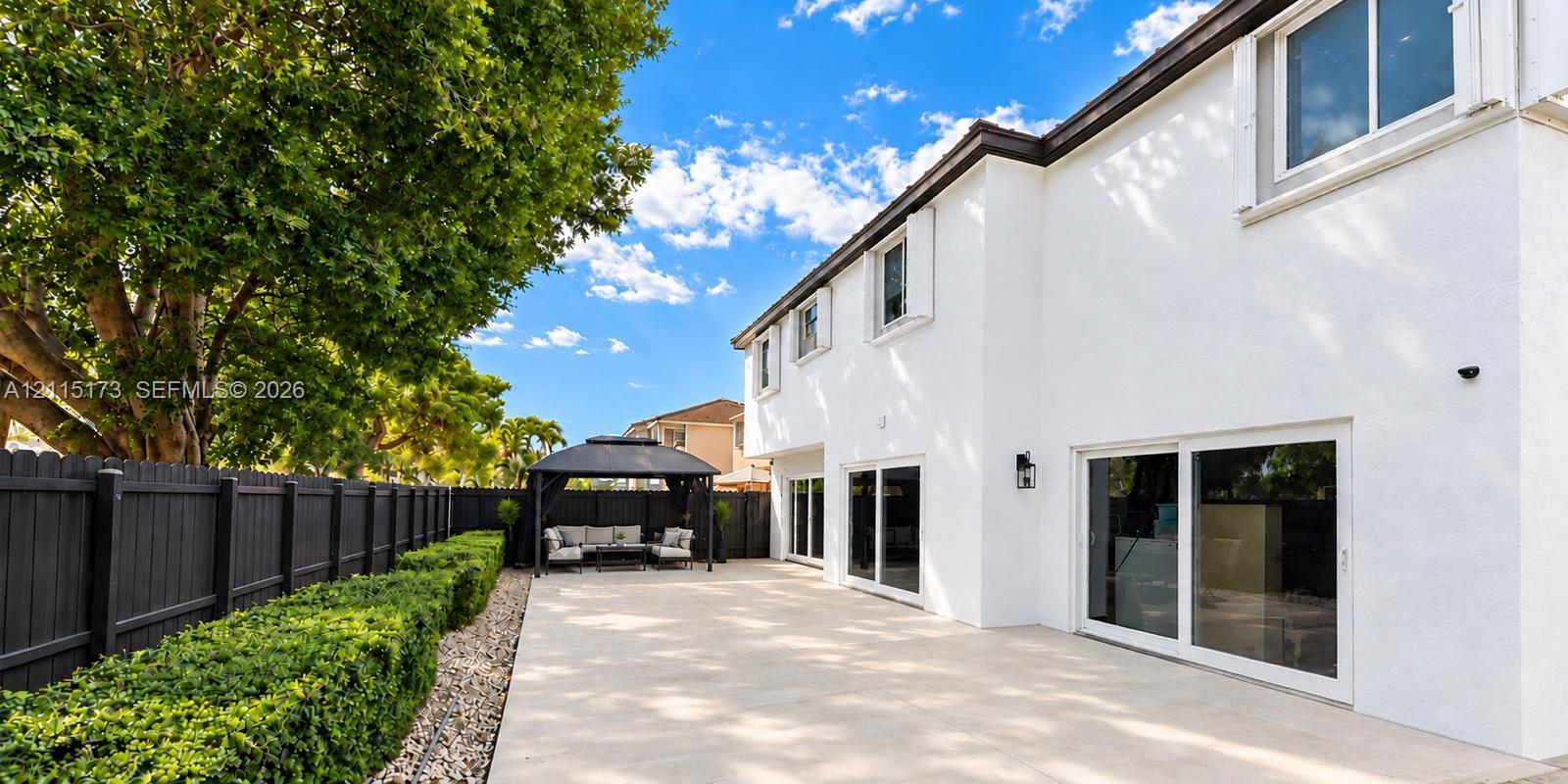Exterior patio featuring expansive light-toned pavers, dark-stained privacy fencing, mature landscaping, and multiple sliding glass door entries