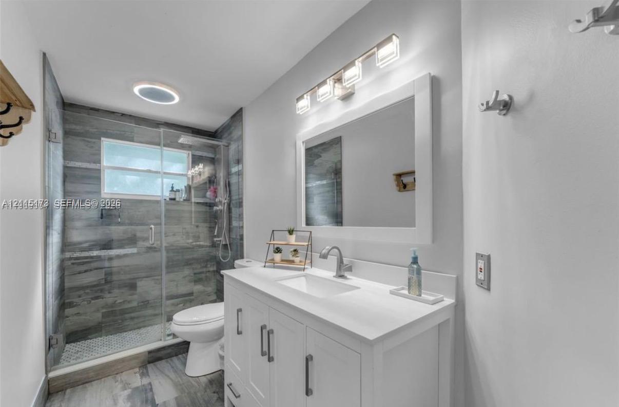 White vanity cabinet with integrated rectangular sink and stone countertop, large framed wall mirror, four-light vanity fixture, and walk-in shower with gray tile surround and glass door