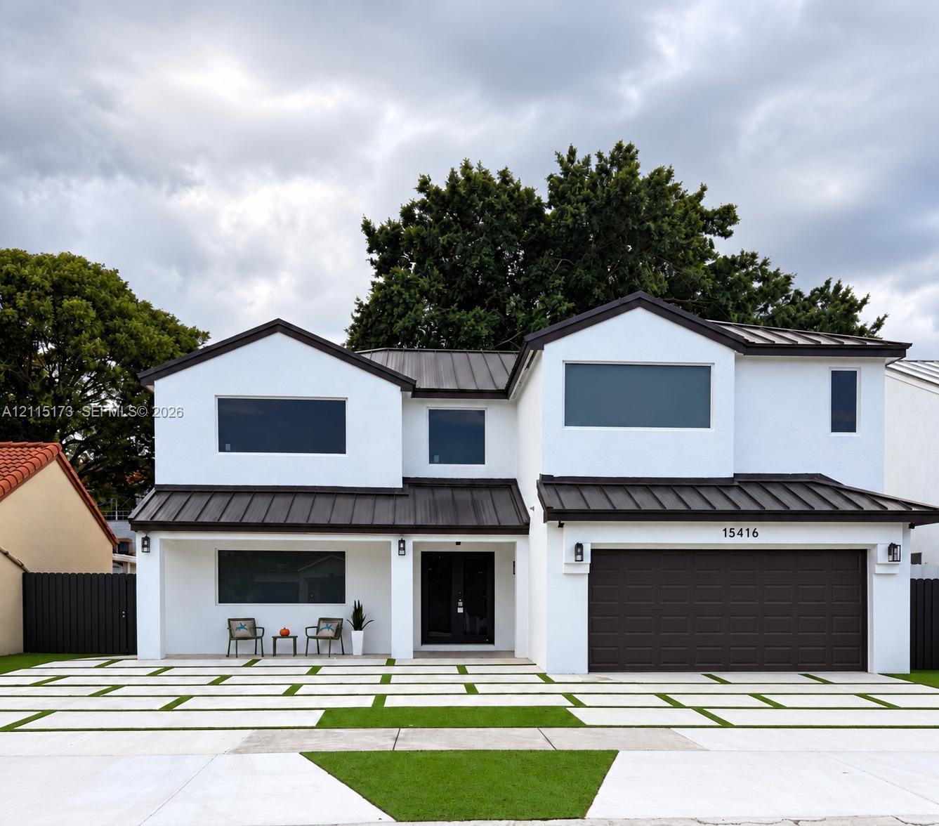 Two-story exterior featuring white stucco finish, dark metal roofing, integrated multi-car garage, and geometric concrete paver driveway with turf accents Two-story exterior featuring white stucco finish, dark metal roofing, integrated multi-car garage, and geometric concrete paver driveway with turf accents