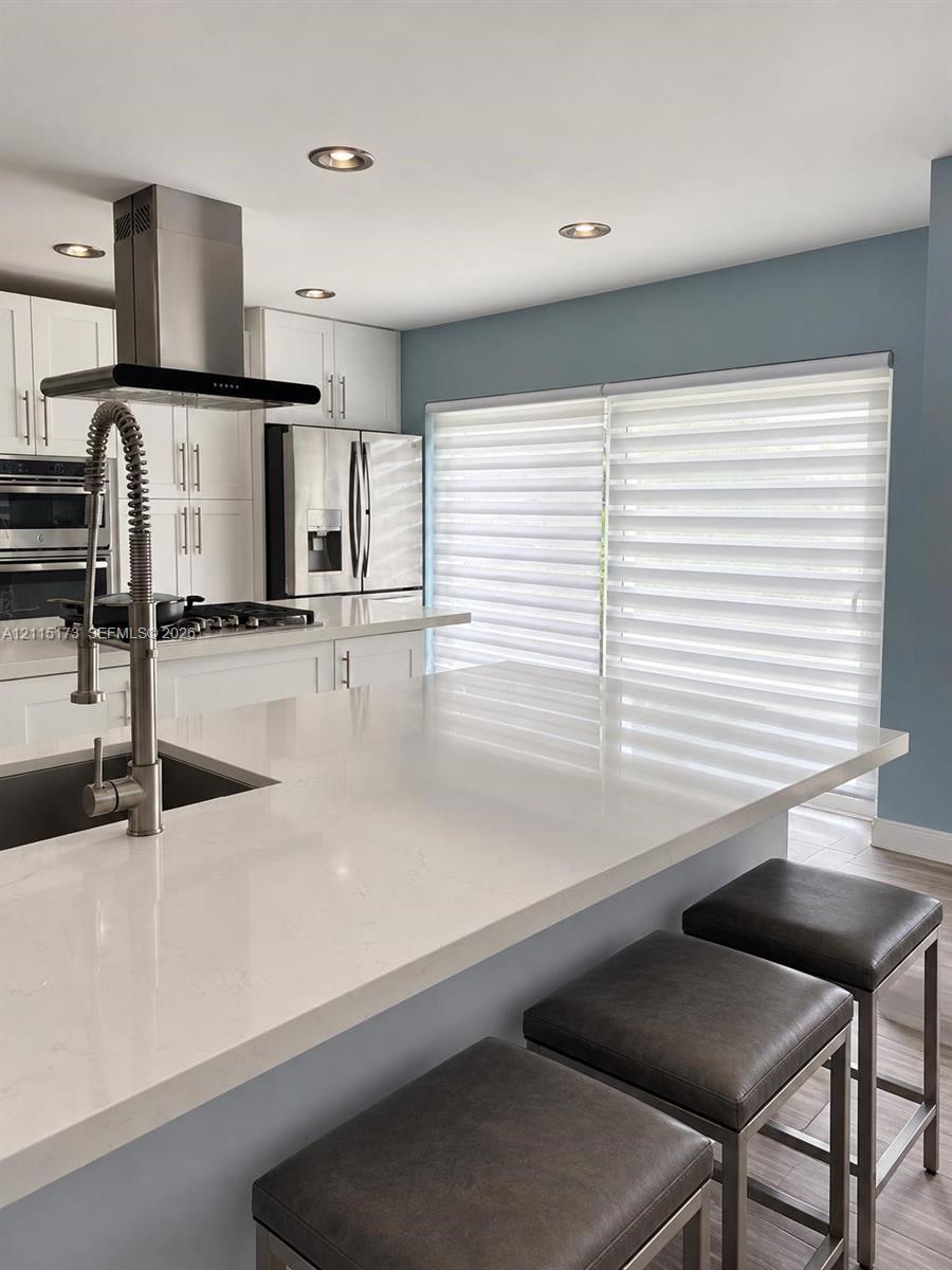 Kitchen featuring a large quartz-topped island with a stainless steel gooseneck faucet, white shaker-style cabinetry, a stainless steel range hood, and recessed ceiling lighting Kitchen featuring a large quartz-topped island with a stainless steel gooseneck faucet, white shaker-style cabinetry, a stainless steel range hood, and recessed ceiling lighting
