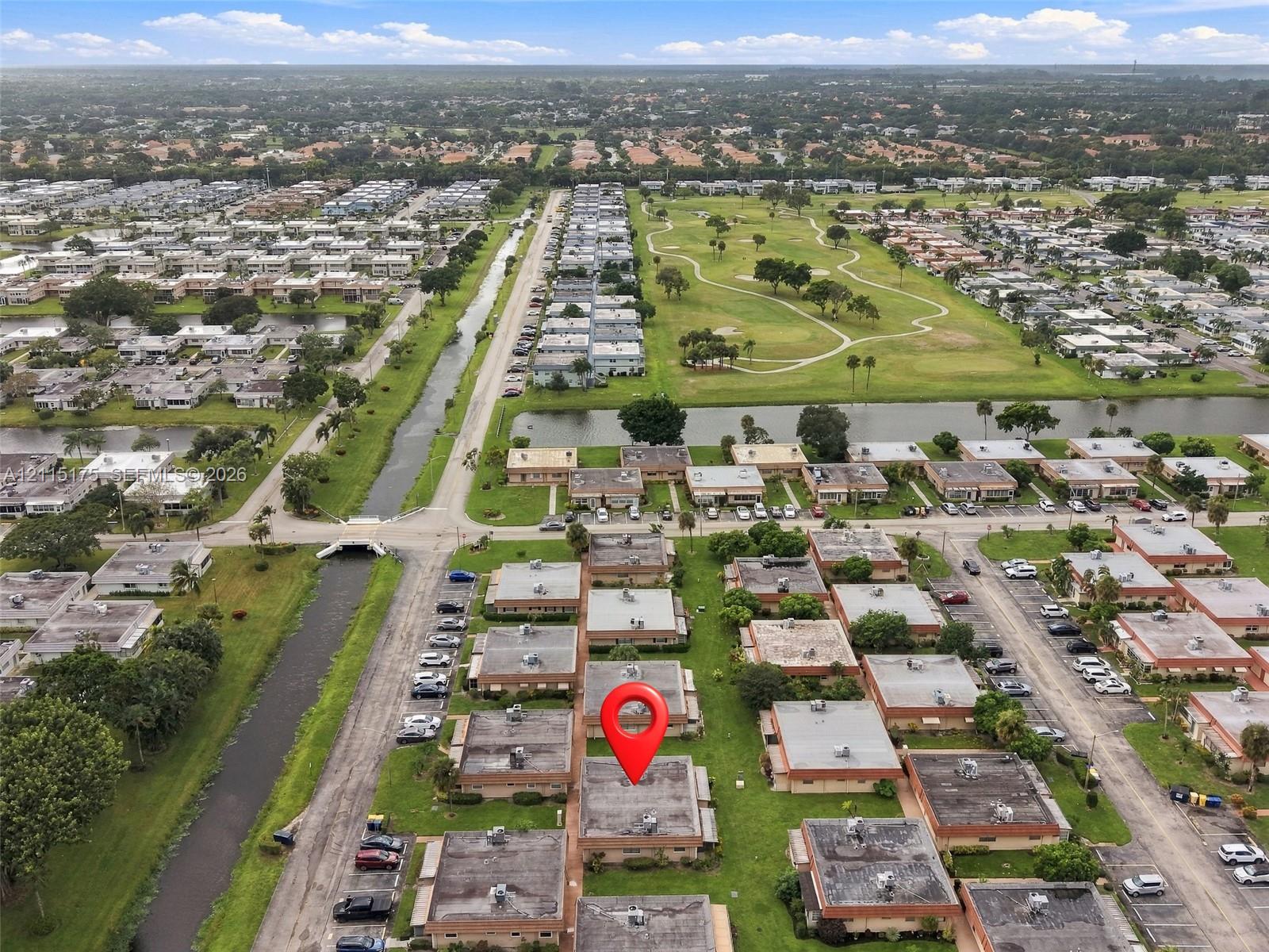 Aerial perspective of a multi-unit residential complex featuring manicured green spaces, water frontage, and proximity to a golf course with a red pin on this listing