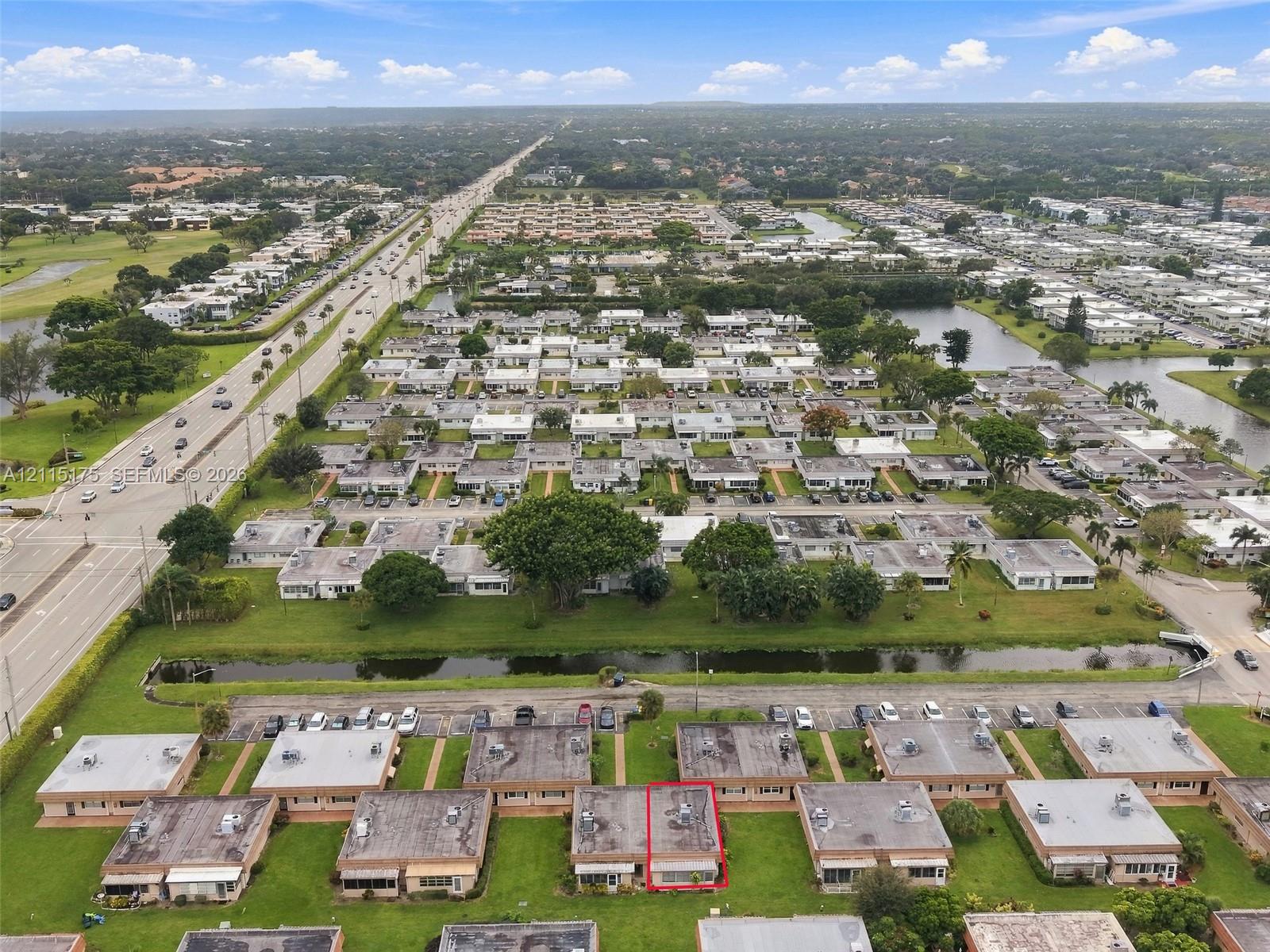 Aerial perspective of a single-story residence featuring a flat roofline, integrated exterior HVAC units, and direct access to a paved parking area, situated within a landscaped neighborhood with water features