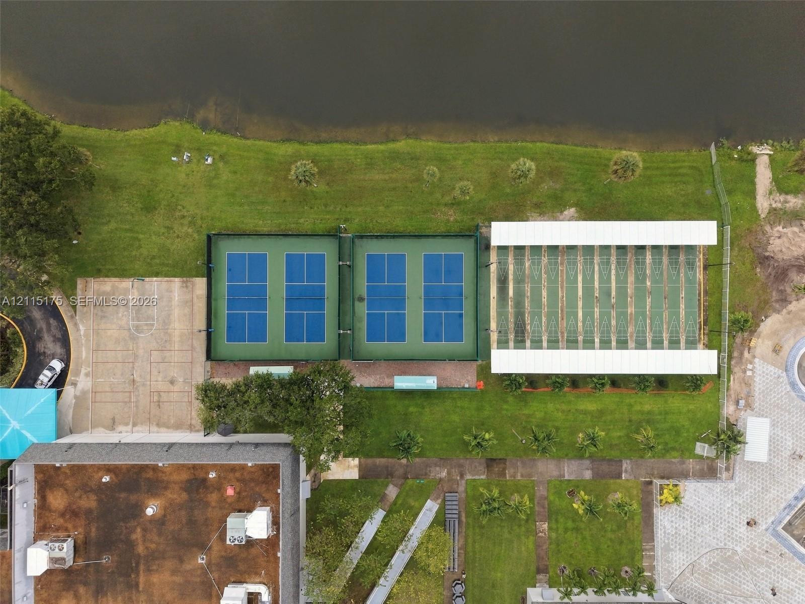 Aerial perspective featuring four fenced pickleball courts, a covered multi-lane shuffleboard area, a concrete sport court, and waterfront grounds
