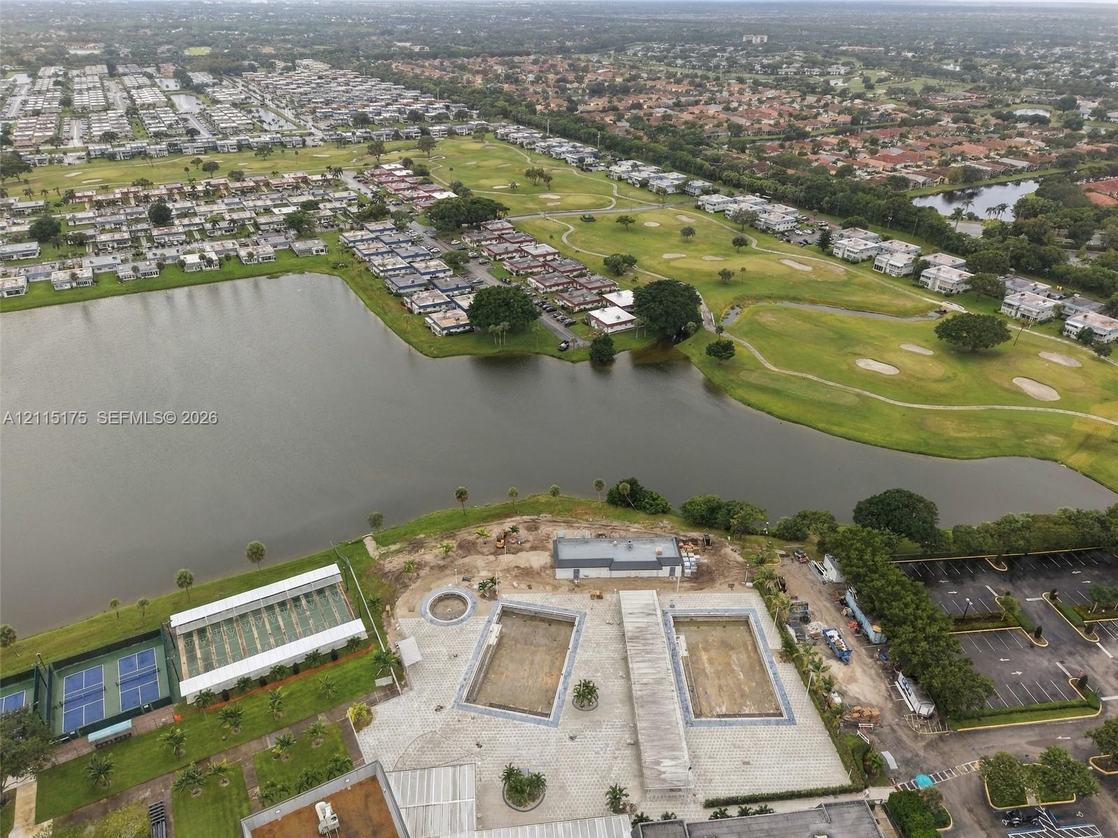 Aerial perspective of the community facilities, featuring two rectangular swimming pools, a dedicated tennis court structure, paved parking area, and waterfront positioning