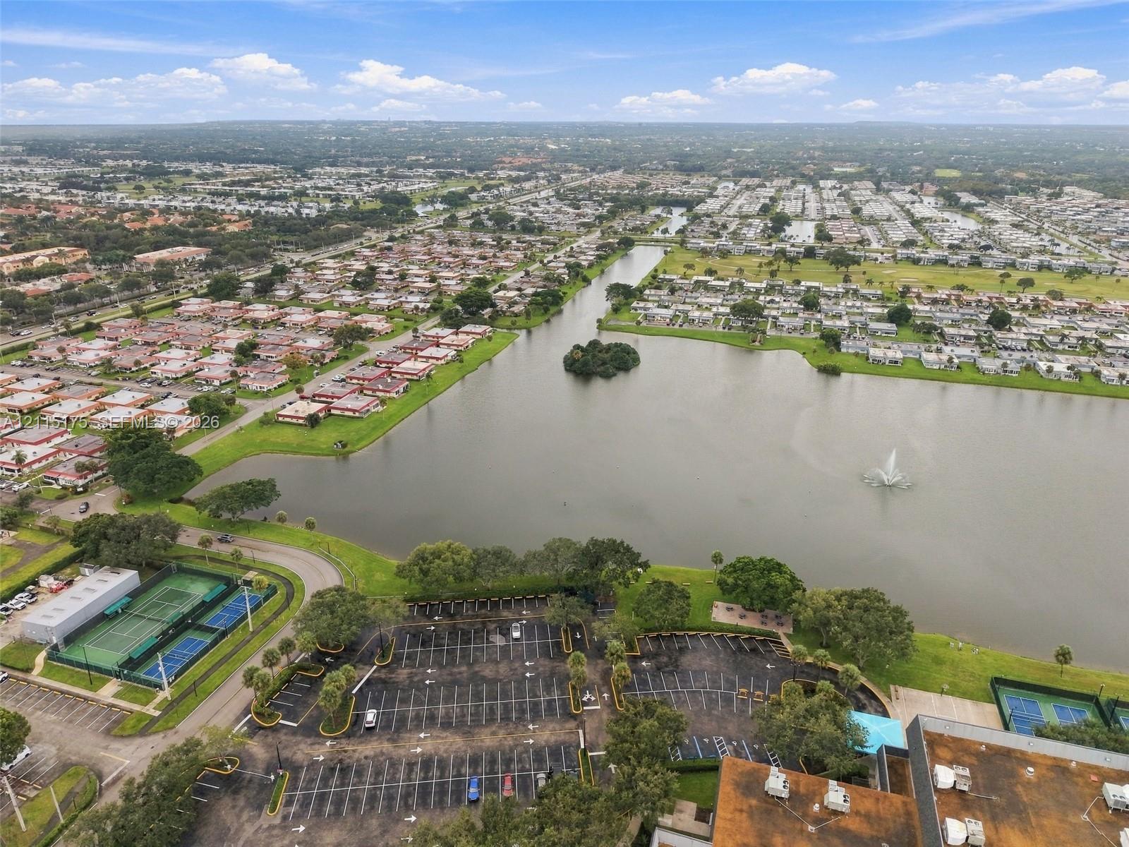Aerial perspective of a lakefront setting featuring a central water fountain, multiple tennis courts, and paved parking areas
