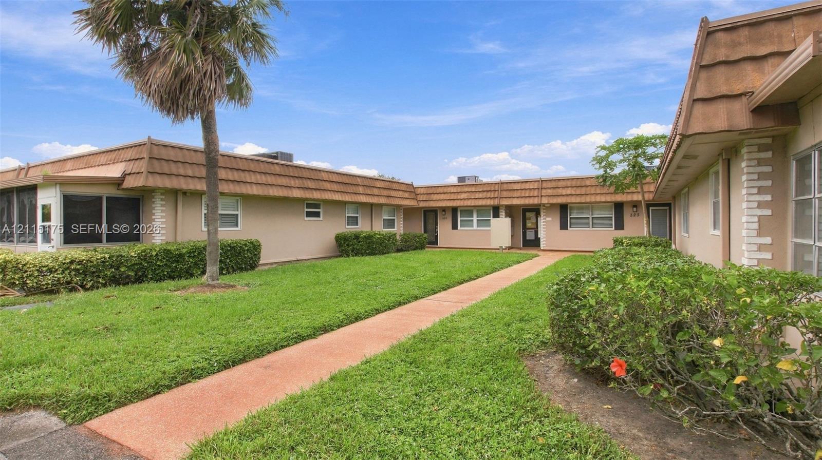Single-story exterior featuring stucco siding, manicured hedges, mature palm trees, and a concrete walkway