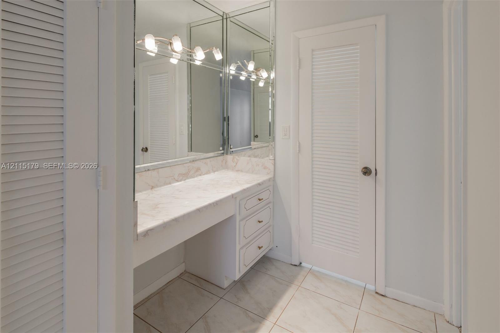 Built-in vanity with stone-surfaced countertop, three-drawer cabinetry, full-height wall mirrors, five-bulb integrated light fixture, and louvered doors