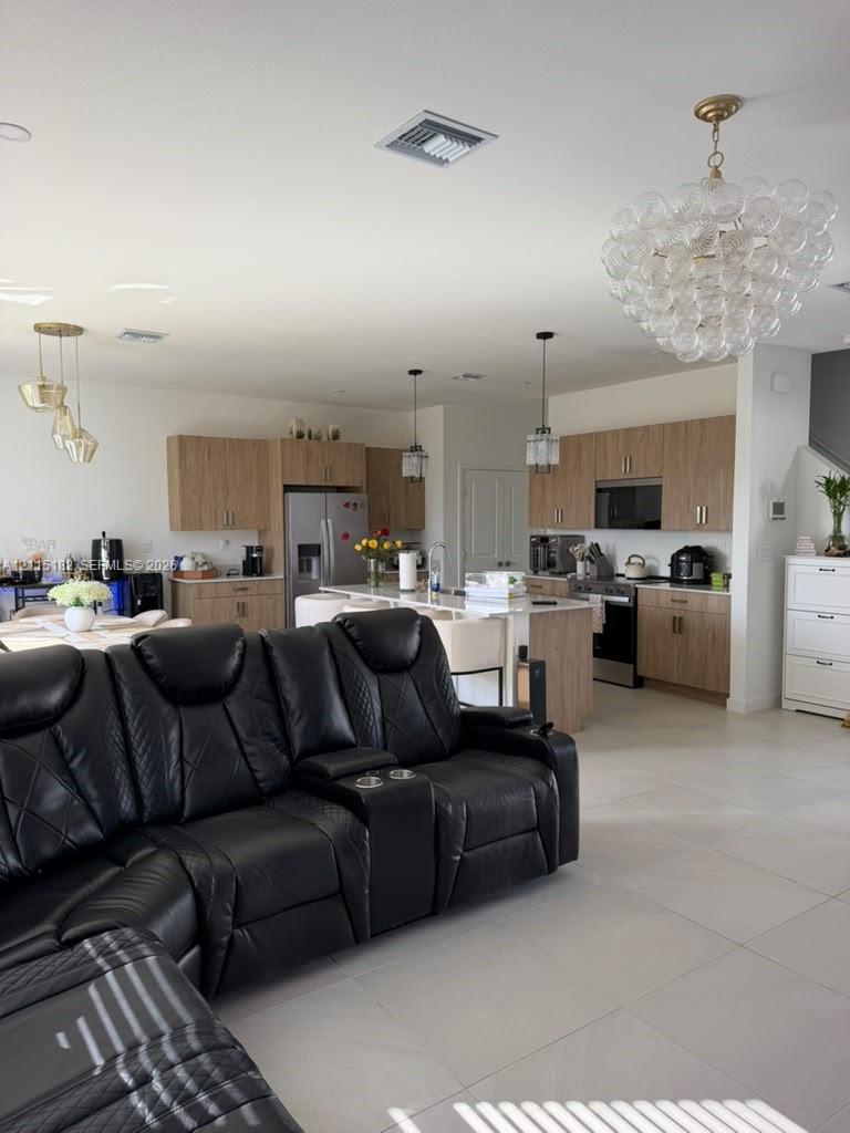 Kitchen featuring wood-finish cabinetry, a large central island with a stone-top counter, stainless steel appliances, large-format tile flooring, and decorative glass chandeliers