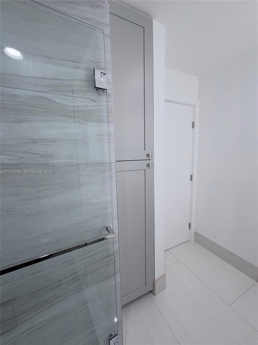Floor-to-ceiling cabinetry with gray shaker-style doors, glass-enclosed shower featuring horizontal stone-patterned tile, and large-format porcelain tile flooring