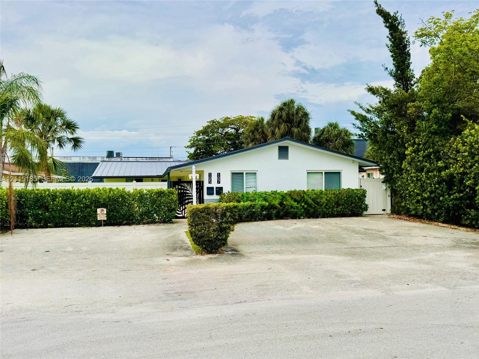 Single-story residence featuring a low-pitch roof, light-colored exterior finish, multi-pane windows, and a paved parking surface enclosed by mature hedge landscaping Single-story residence featuring a low-pitch roof, light-colored exterior finish, multi-pane windows, and a paved parking surface enclosed by mature hedge landscaping