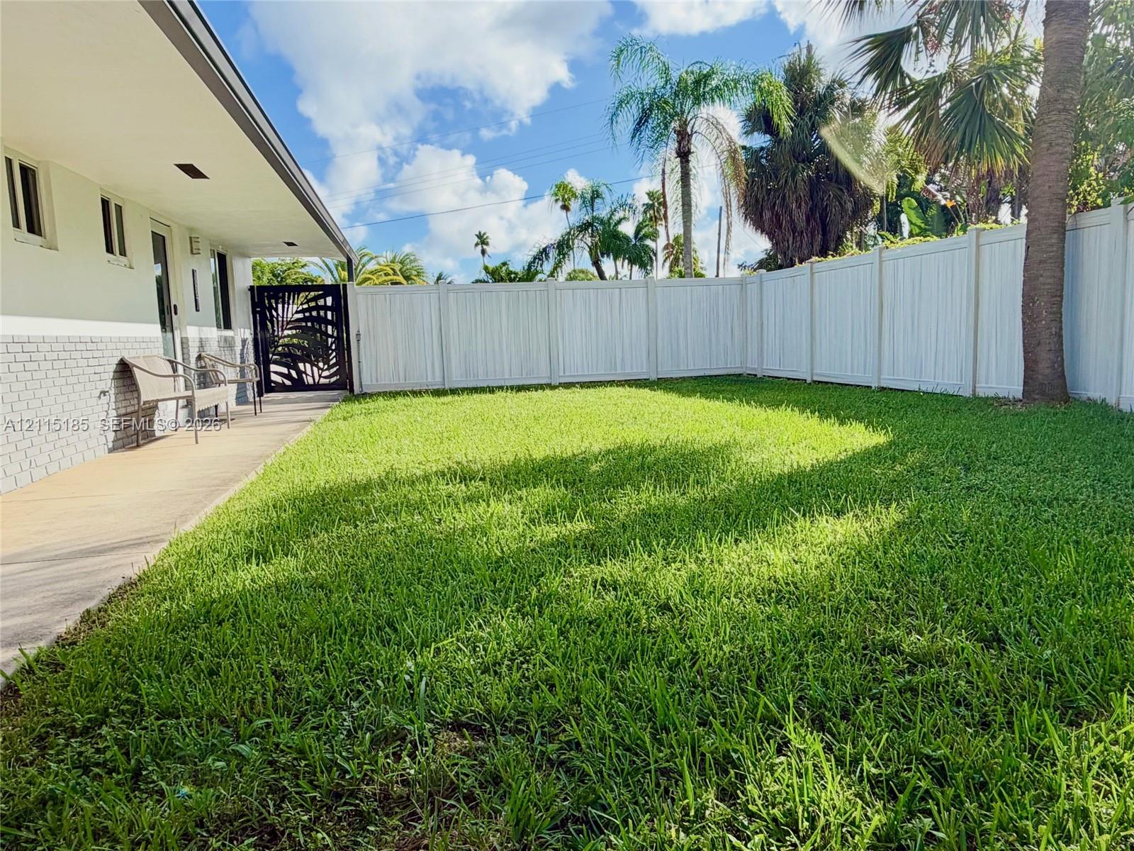 Backyard featuring a paved covered patio, manicured lawn, white vinyl fencing, and an ornate metal gate Backyard featuring a paved covered patio, manicured lawn, white vinyl fencing, and an ornate metal gate