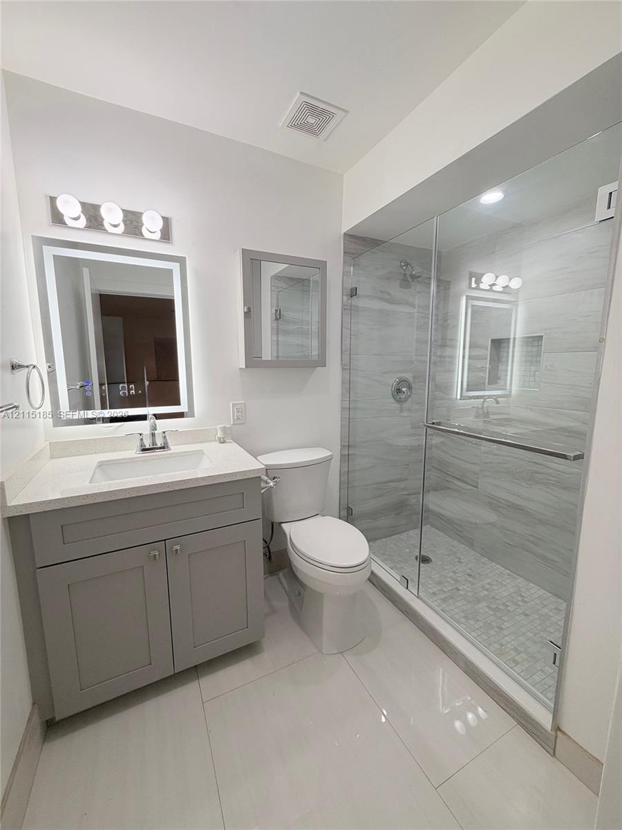Bathroom featuring a vanity with a rectangular sink and stone-finish countertop, backlit mirror, grey cabinetry, and a glass-enclosed shower with tiled walls and flooring