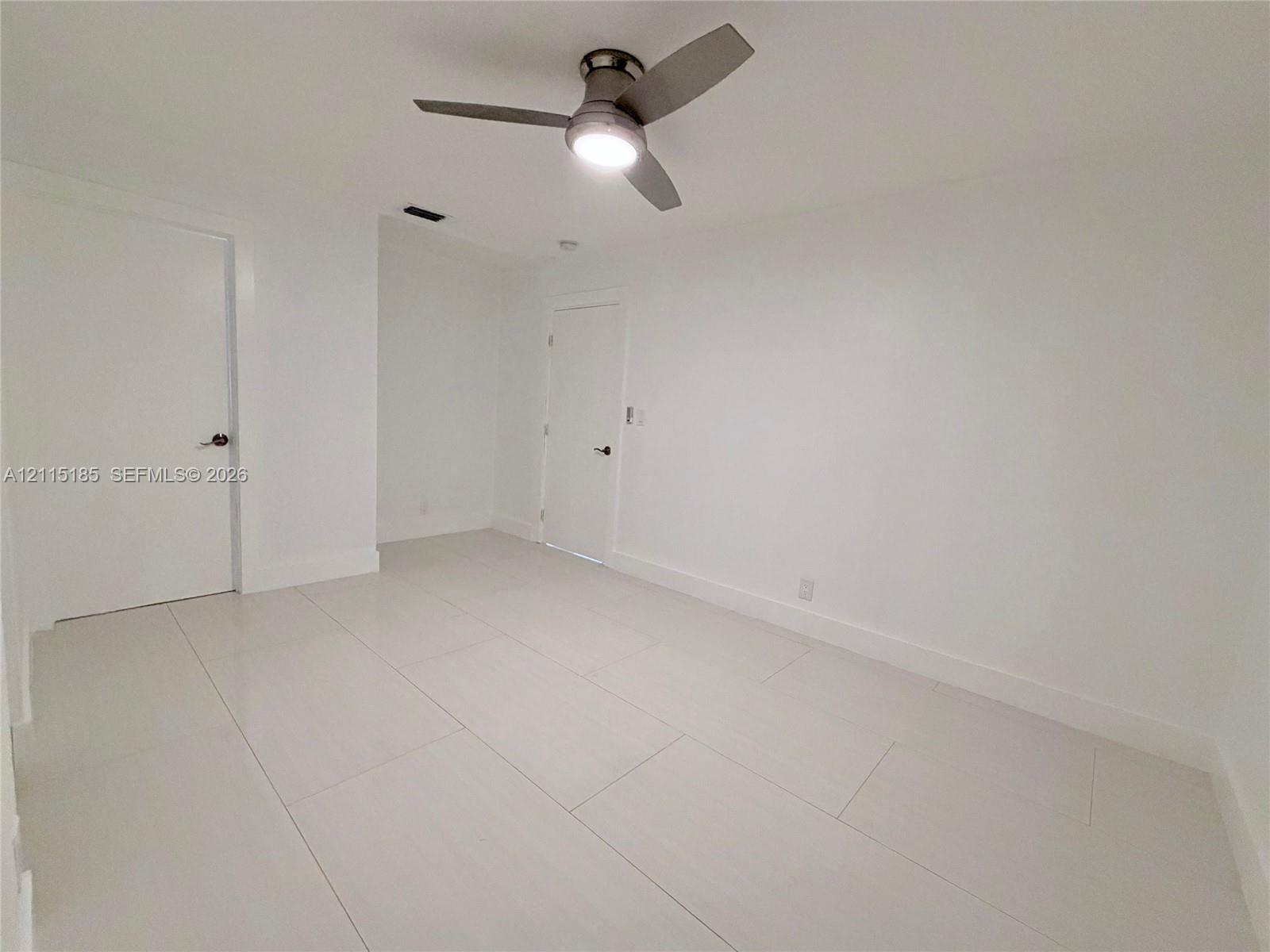 Interior space featuring large-format porcelain tile flooring, smooth wall finishes, two interior doors with matte-finish hardware, and a modern three-blade ceiling fan with integrated lighting