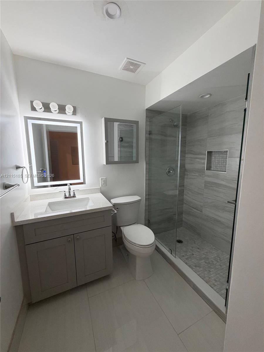 Bathroom featuring a vanity with white countertop, integrated sink, and illuminated mirror, paired with a walk-in shower encased in glass with stone-finish wall tiling and a built-in recessed niche