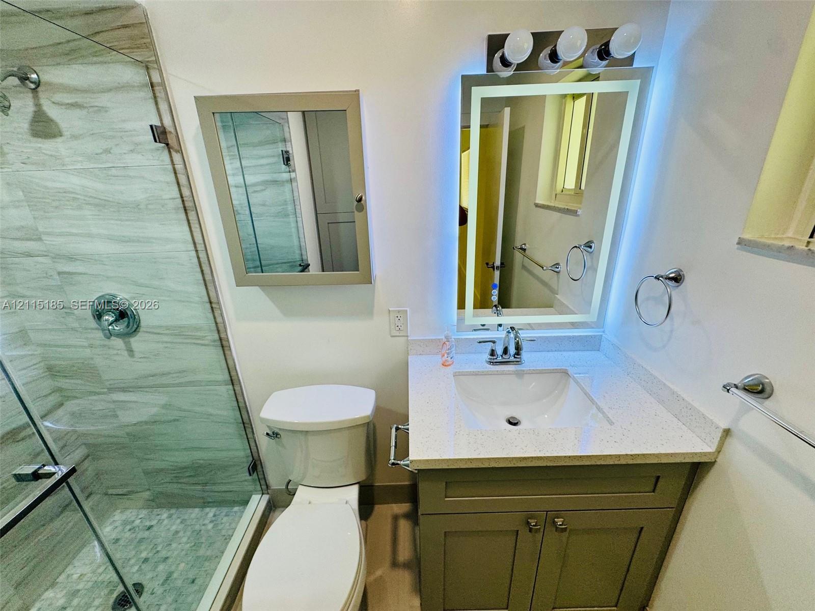 Bathroom featuring a vanity with a light-toned countertop, integrated basin, illuminated mirror with integrated lighting, a walk-in shower with stone-finish tile surround and pebble tile flooring, and a glass shower enclosure