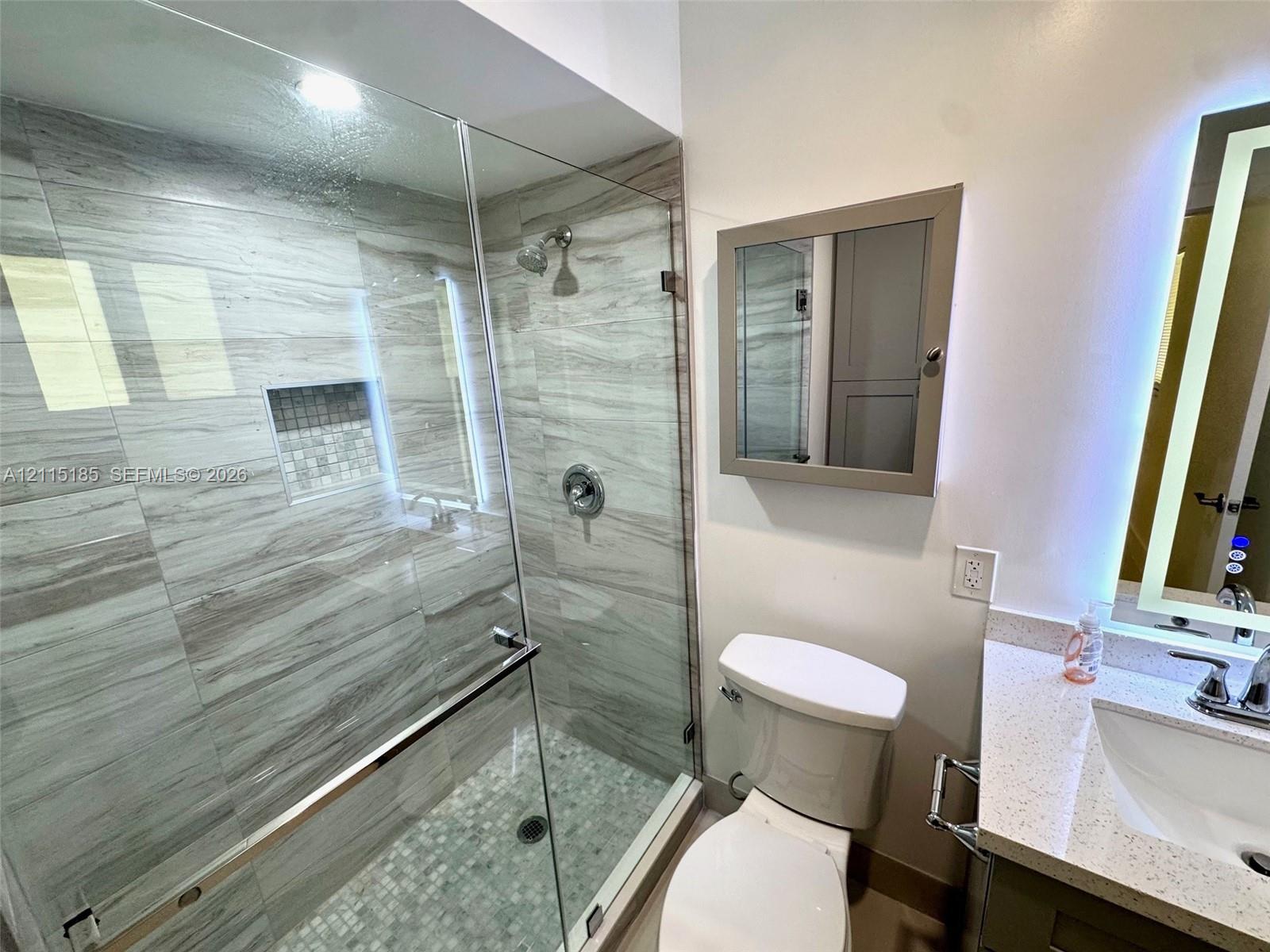 Glass-enclosed shower featuring large-format tile surround, integrated tiled niche, stone-composite vanity countertop, and backlit vanity mirror