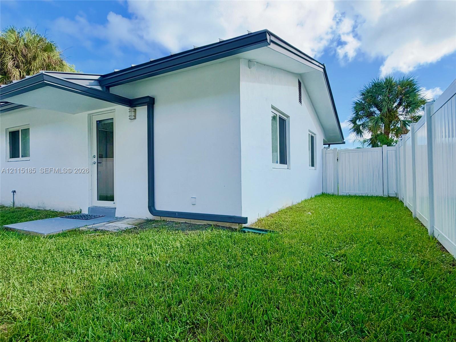 Exterior facade featuring white stucco finish, dark-trimmed eaves, a glass-paneled exterior door, and a white vinyl privacy fence enclosing the lawn