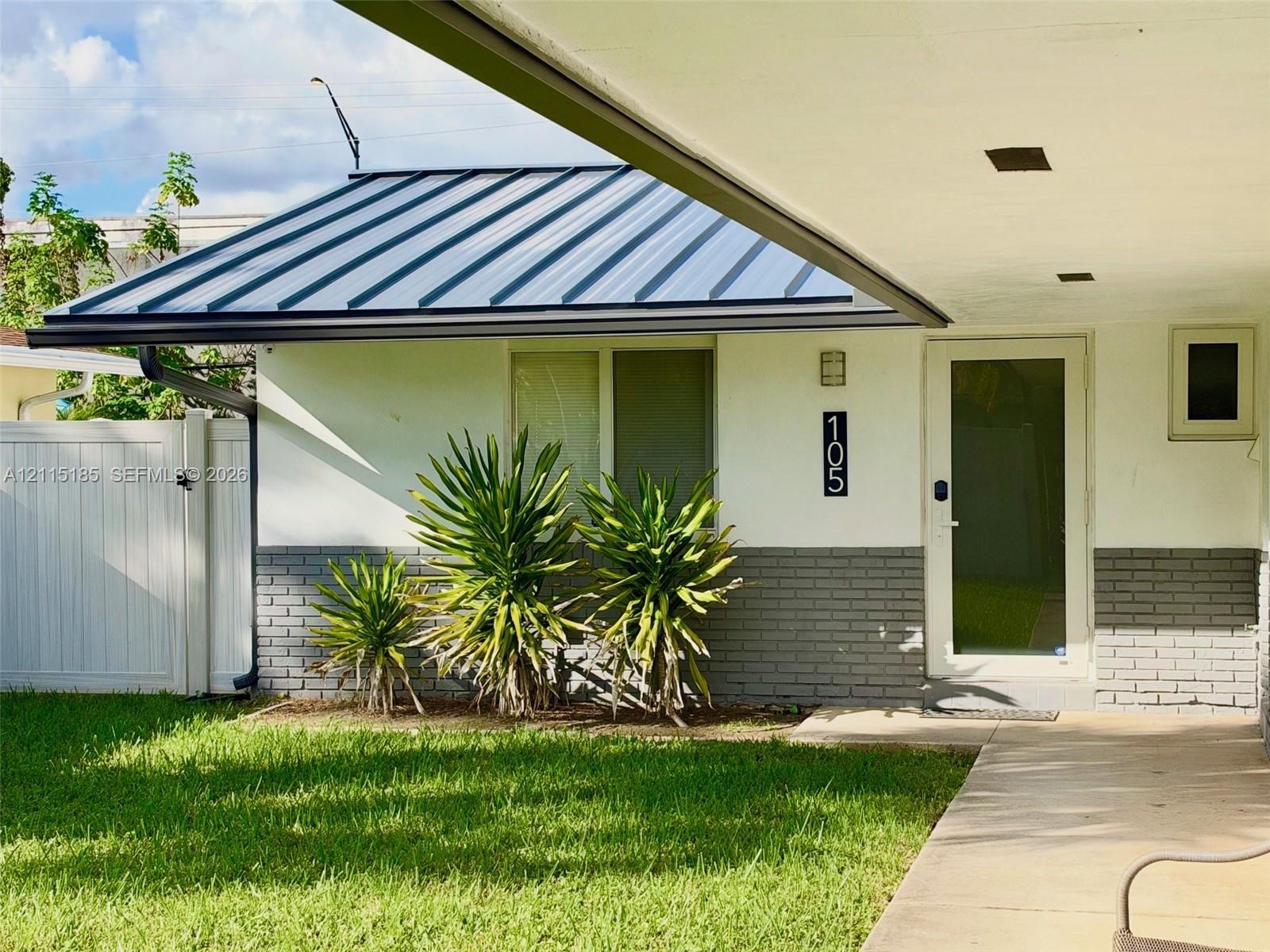 Exterior featuring a standing-seam metal roof, white stucco walls with gray brick veneer, a covered entryway, and a white privacy fence Exterior featuring a standing-seam metal roof, white stucco walls with gray brick veneer, a covered entryway, and a white privacy fence