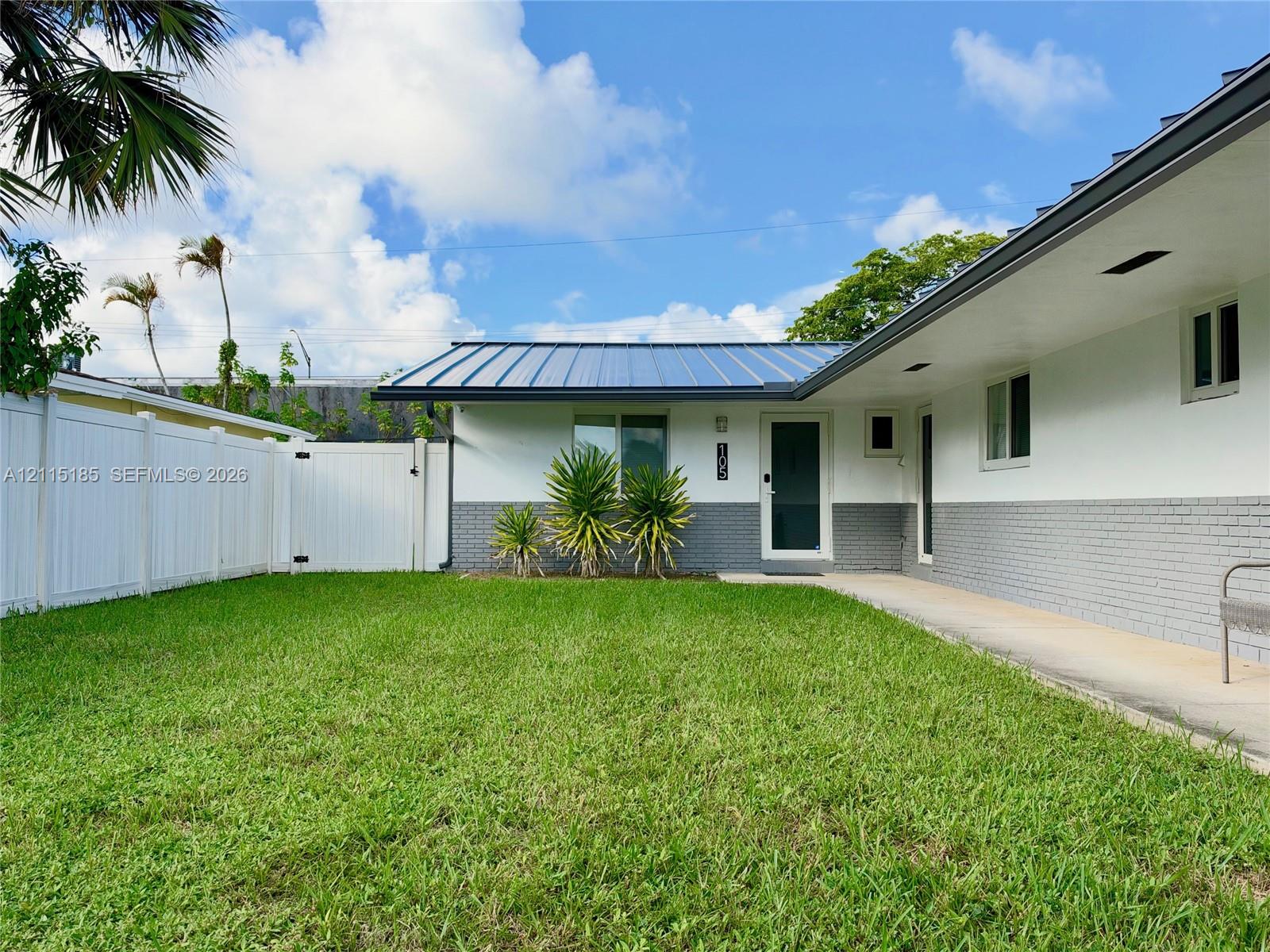 Exterior facade featuring a standing-seam metal roof, white stucco walls with grey brick lower siding, and a white vinyl privacy fence enclosing a grass lawn