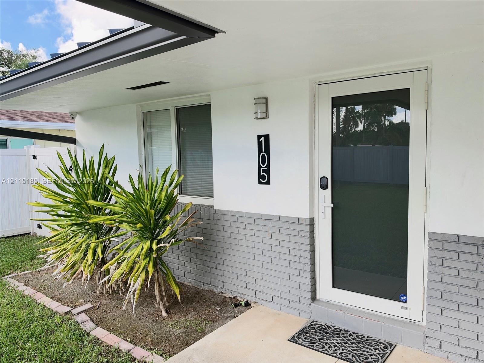 Exterior entrance featuring a white-painted finish with a gray brick wainscot, a full-view storm door, a covered porch, and a brick-edged landscape bed