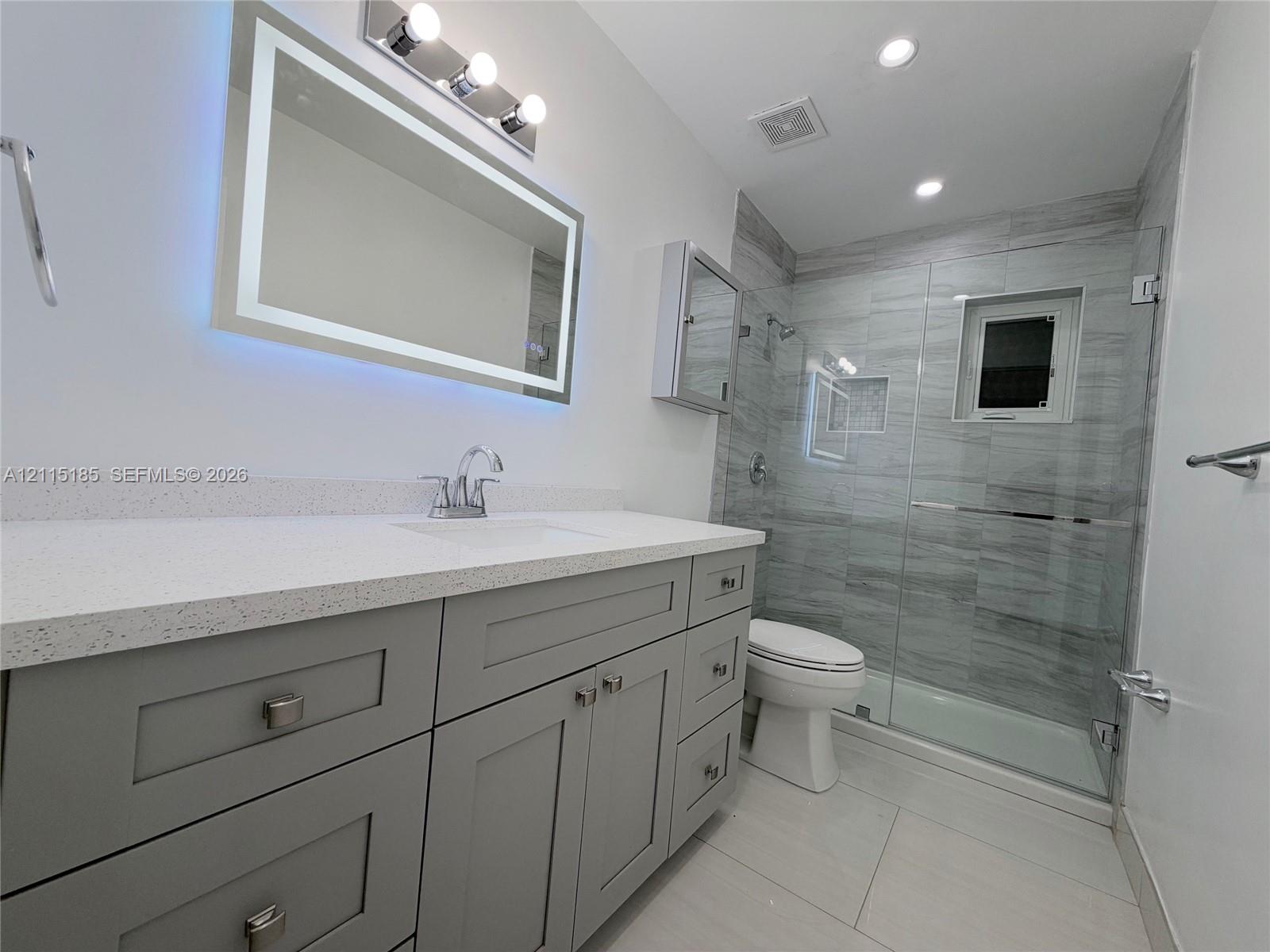 Grey shaker-style vanity with a light-toned solid surface countertop, rectangular undermount sink, illuminated mirror, and glass-enclosed shower featuring horizontal-pattern wall tiling