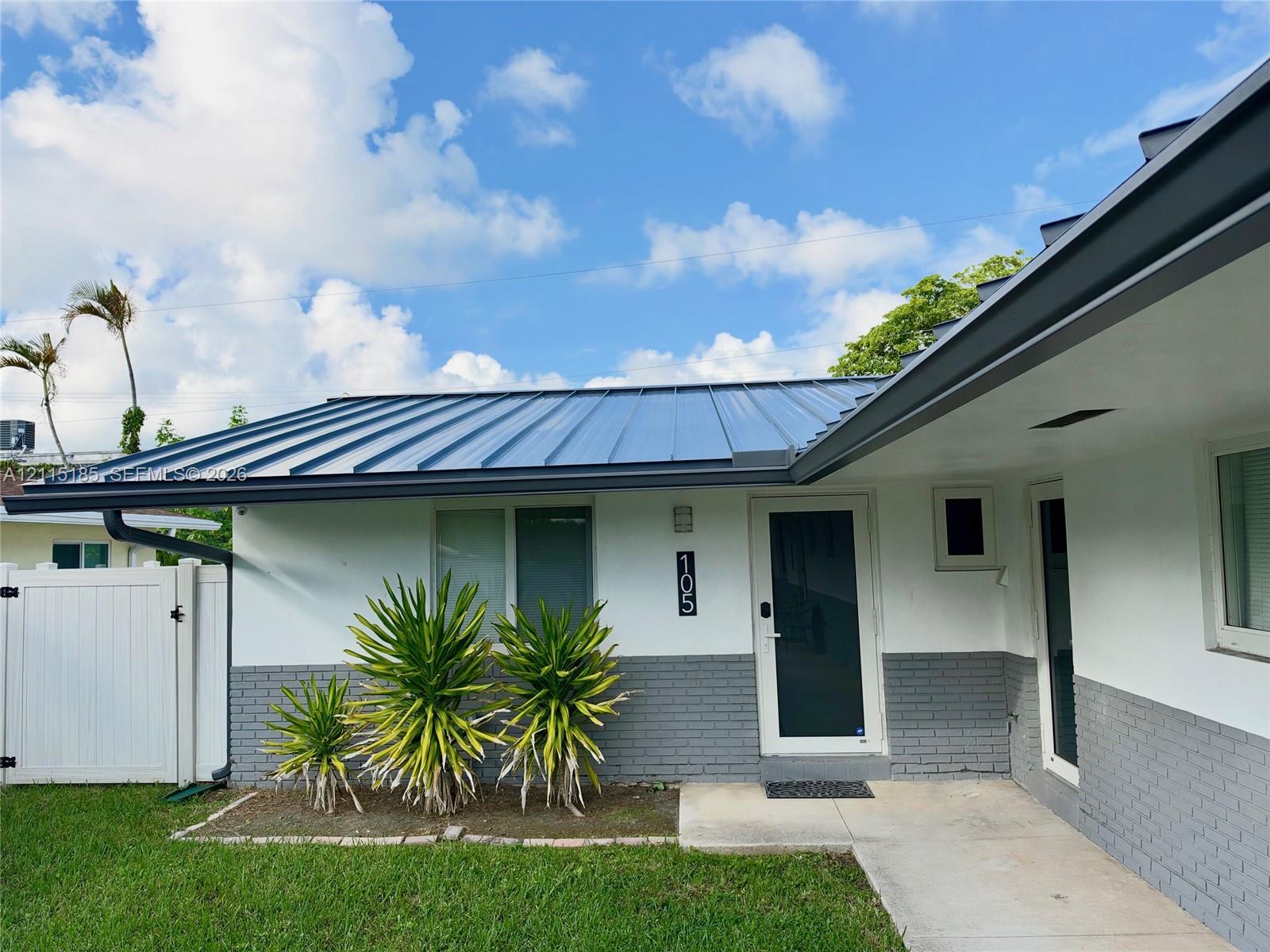 Exterior facade featuring a metal roof, two-tone painted brick and smooth finish siding, concrete entryway, and white vinyl privacy fencing