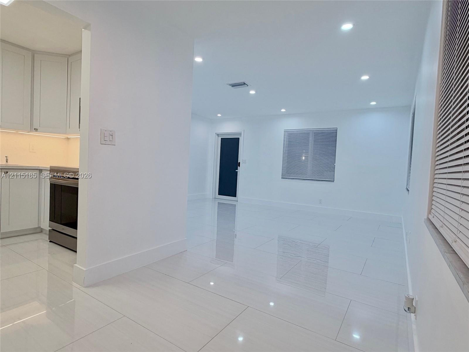 Open interior featuring large-format ceramic tile flooring, recessed ceiling lighting, white shaker-style cabinetry, and a dark-toned door