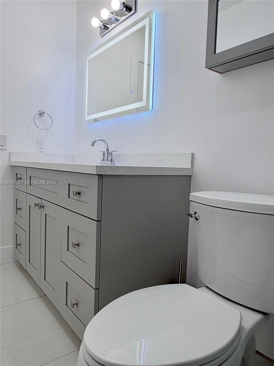 Gray shaker-style vanity with stone-composite countertop, illuminated LED vanity mirror, chrome faucet, and tiled flooring