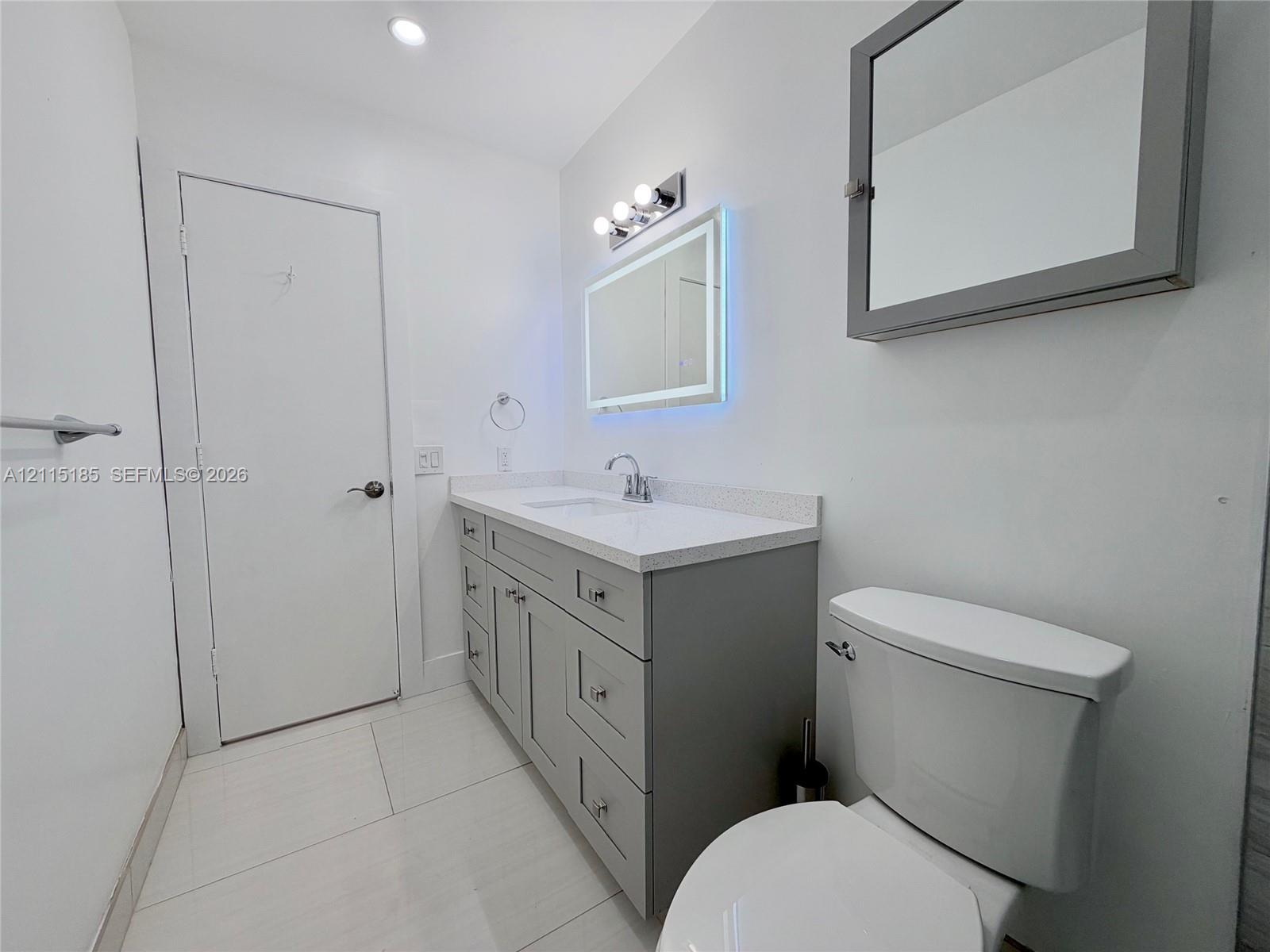 Bathroom featuring a gray vanity with a stone countertop, an integrated sink, an illuminated vanity mirror, and large-format tile flooring