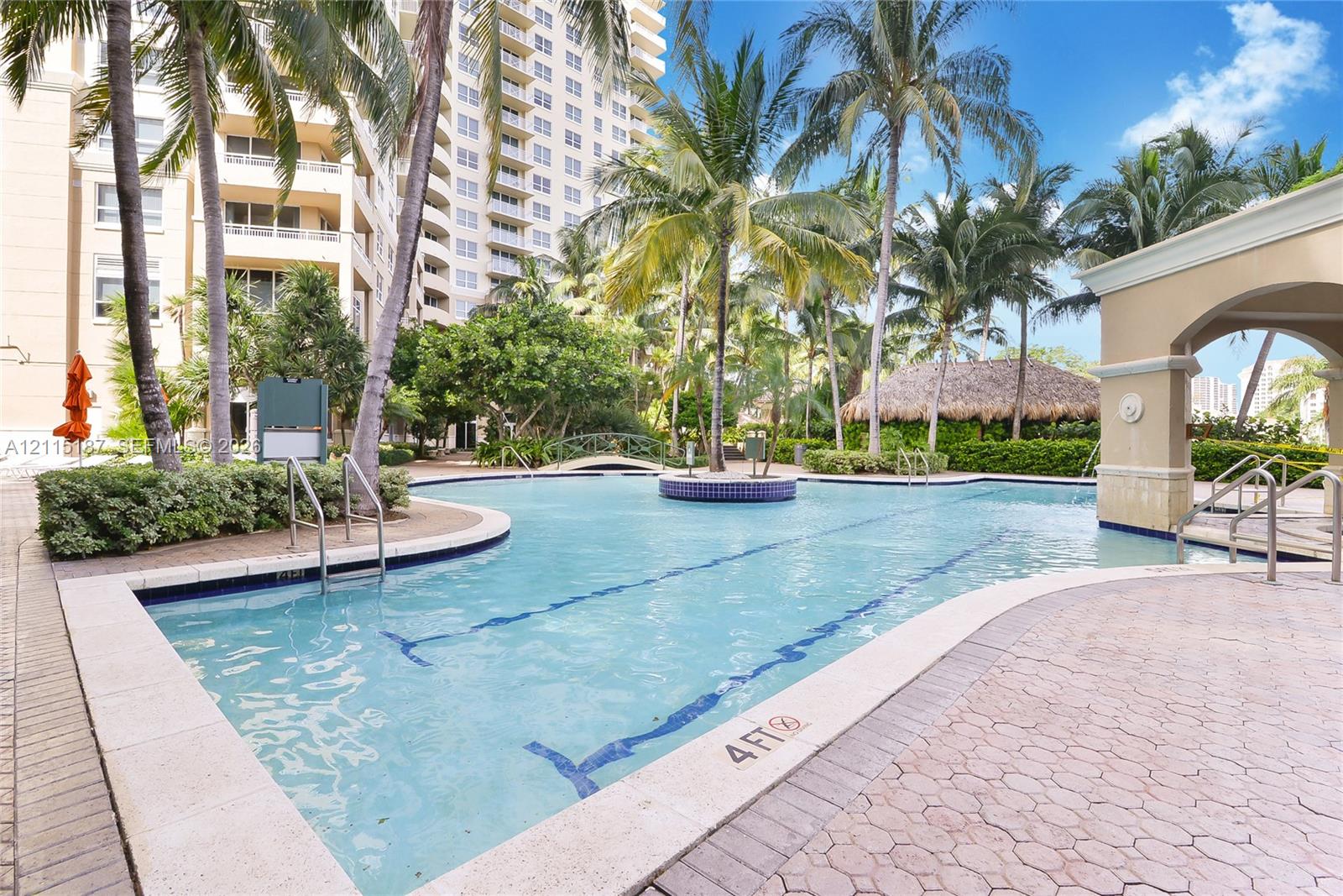 Swimming pool with a paved deck, surrounding palm trees, thatched-roof structure, and arched architectural detailing