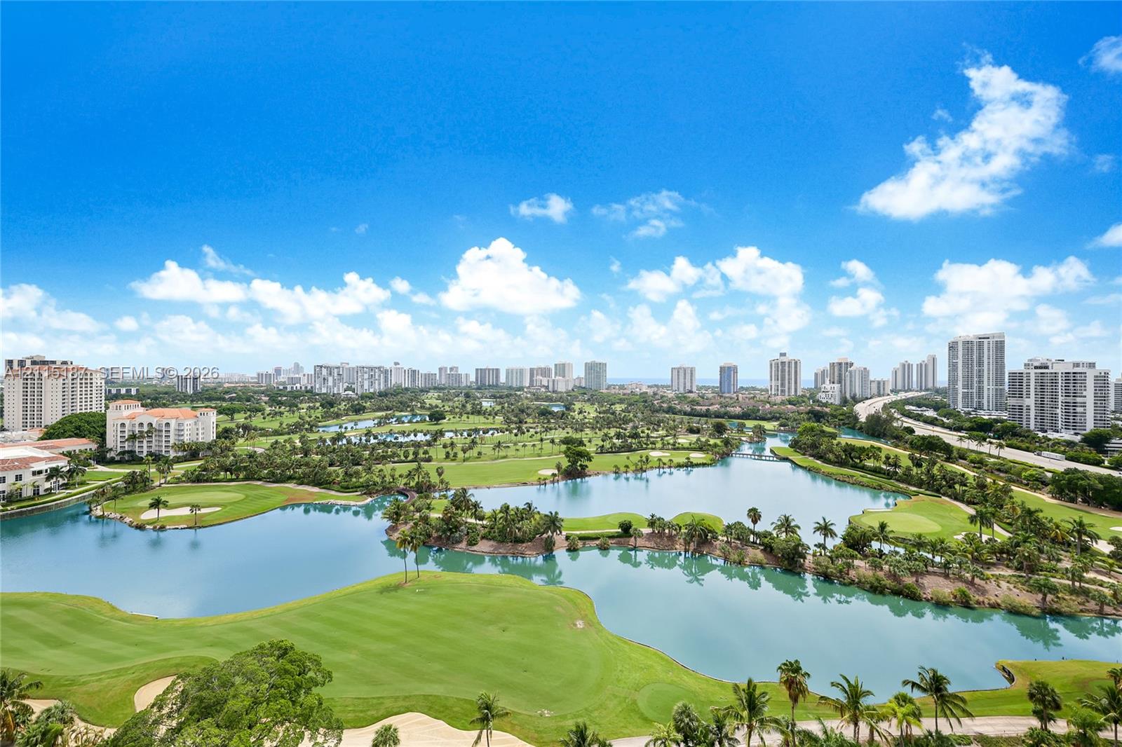 Panoramic perspective featuring a golf course, expansive water features, mature palm trees, and an urban skyline