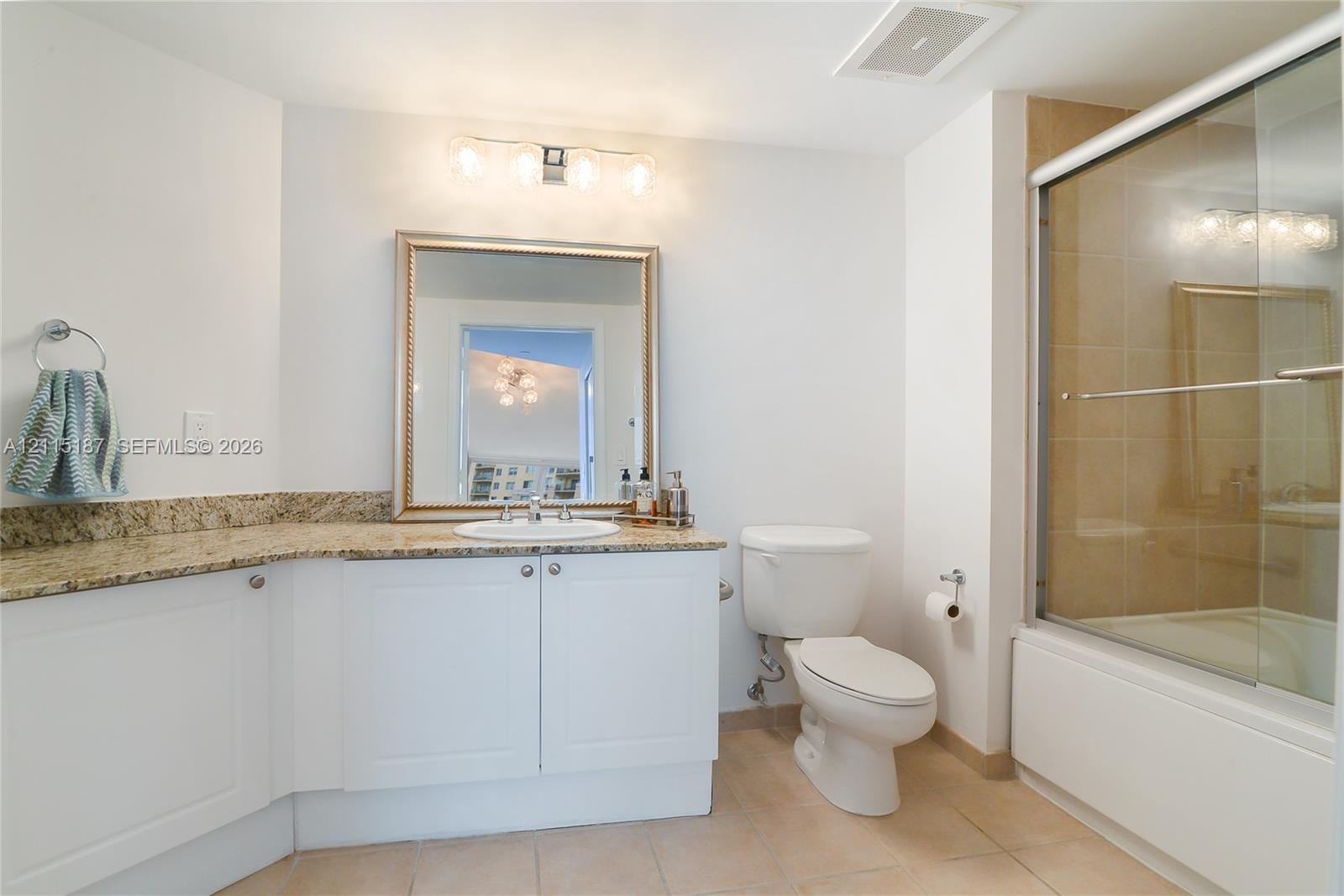 Bathroom featuring white cabinetry, stone-surface countertop with an integrated basin, framed wall mirror, four-light vanity fixture, and a shower-tub combination with sliding glass doors Bathroom featuring white cabinetry, stone-surface countertop with an integrated basin, framed wall mirror, four-light vanity fixture, and a shower-tub combination with sliding glass doors