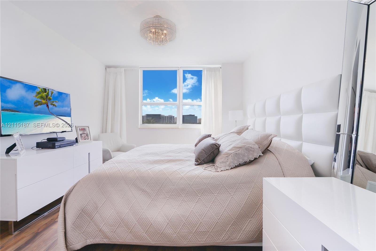 Bedroom featuring wood-finish flooring, a large multi-pane window, and a decorative crystal-style flush-mount ceiling light fixture Bedroom featuring wood-finish flooring, a large multi-pane window, and a decorative crystal-style flush-mount ceiling light fixture