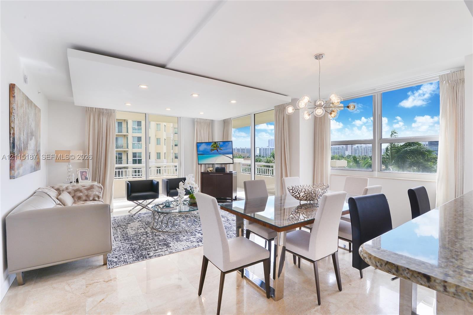 Open-concept interior featuring large-format stone flooring, floor-to-ceiling windows, a dropped tray ceiling with recessed lighting, and an expansive multi-light chandelier Open-concept interior featuring large-format stone flooring, floor-to-ceiling windows, a dropped tray ceiling with recessed lighting, and an expansive multi-light chandelier
