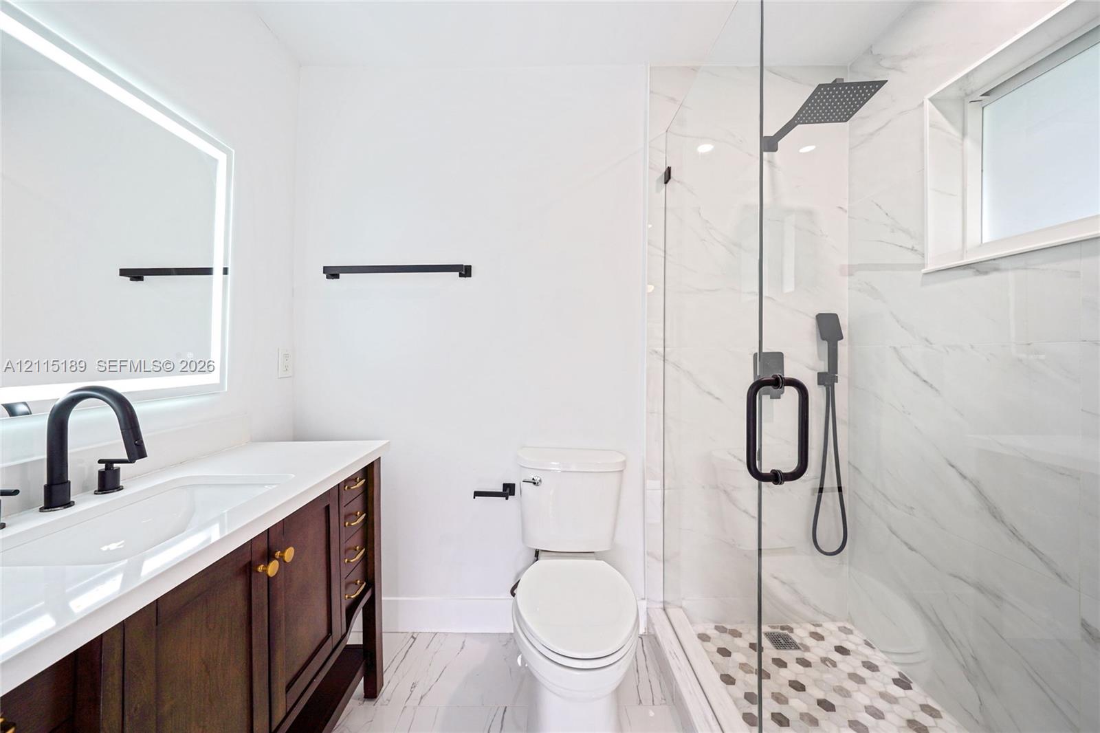 Bathroom featuring a dark wood vanity with a white countertop, backlit vanity mirror, walk-in shower with matte black fixtures, and patterned shower floor tile