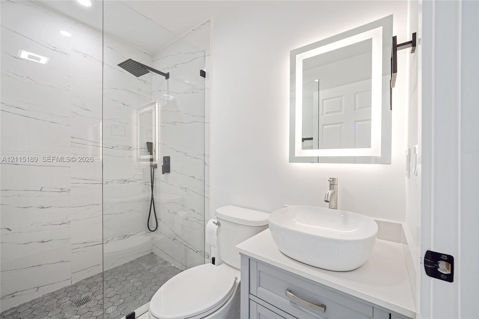 Bathroom featuring a walk-in shower with large-format stone-patterned tile, a frameless glass enclosure, a vessel sink on a vanity with a solid-surface countertop, and an integrated LED-backlit mirror