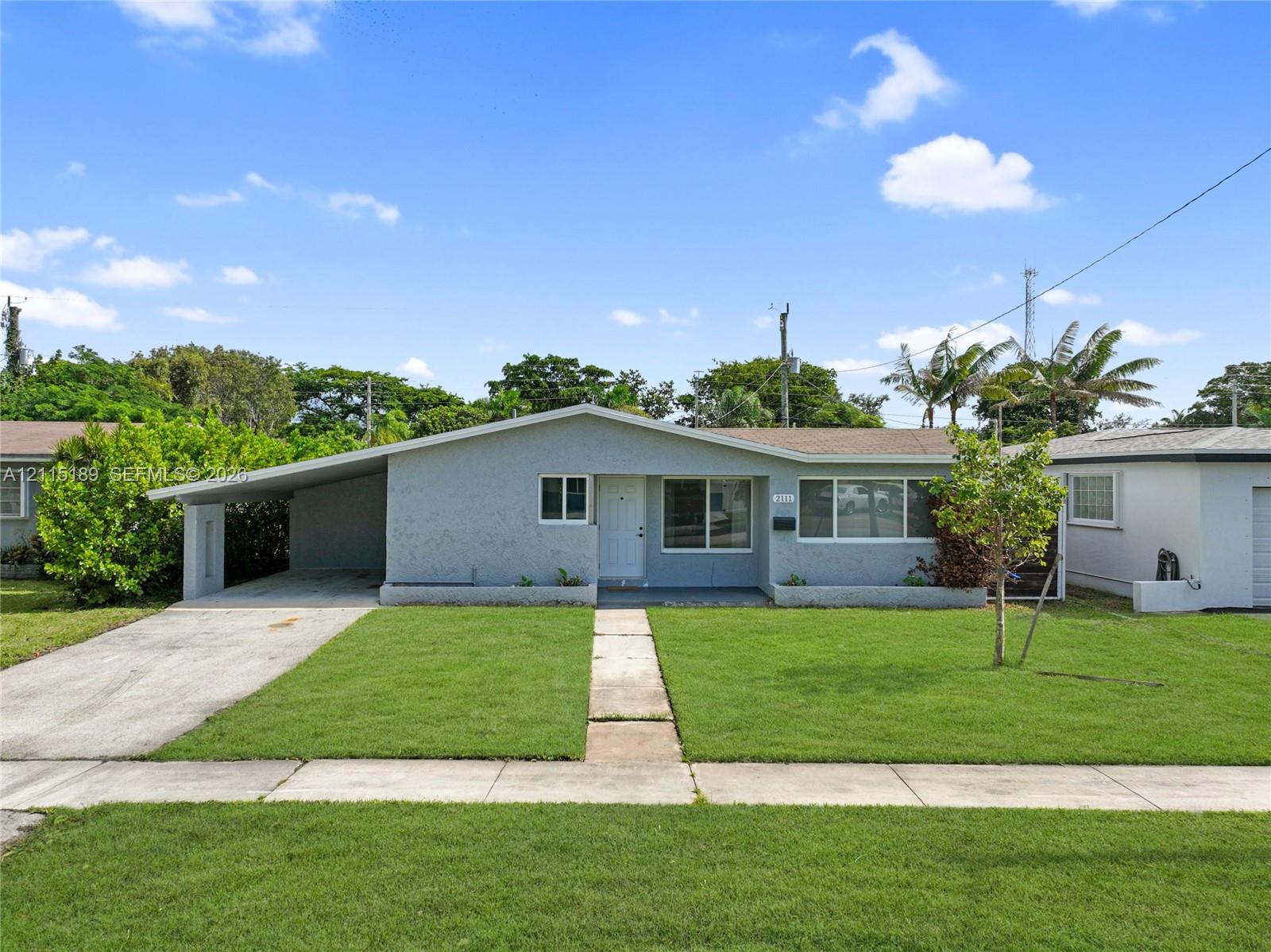 Single-story exterior featuring a light gray stucco finish, an attached carport, a concrete driveway, and a front lawn with established landscaping Single-story exterior featuring a light gray stucco finish, an attached carport, a concrete driveway, and a front lawn with established landscaping