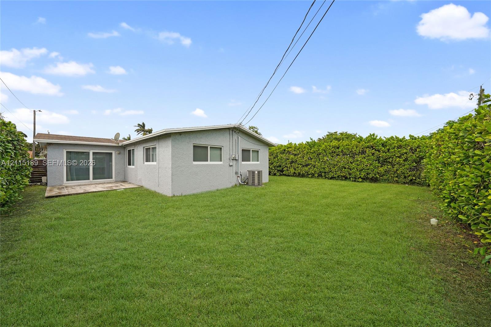 Spacious backyard featuring a grass lawn, concrete patio, sliding glass door, and mature privacy hedging