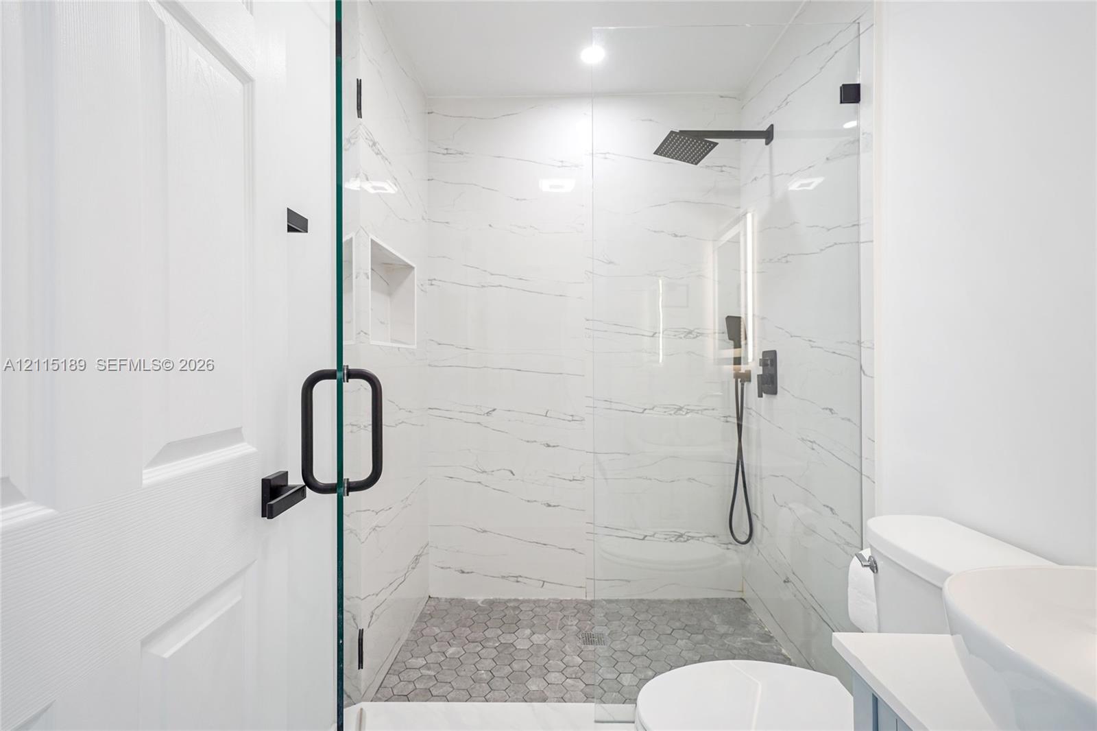 Walk-in shower featuring large-format wall tile, a frameless glass enclosure, a built-in recessed niche, a rainfall showerhead with matte black finish, and a hexagonal tile shower floor